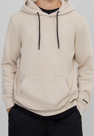 Beige hættetrøje med sorte snore og lomme foran, båret over en sort skjorte, kombineret med sorte bukser.