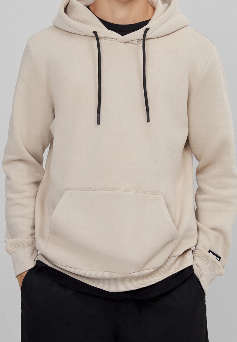 Sweat à capuche beige avec cordons noirs et poche avant, porté sur un t-shirt noir, associé à un pantalon noir.