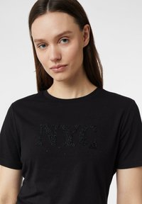 Młoda kobieta z prostymi brązowymi włosami nosi czarną koszulkę z czarnym koralikowym napisem "NYC" na jednolitym jasnym tle.