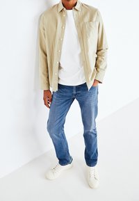 Světlá béžová košile na knoflíky přes bílé tričko, doplněná modrými džínami a bílými teniskami. Košile má náprsní kapsu a měkkou texturu.