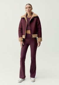 Prošívaná vínová bunda s béžovým umělým kožešinovým límcem a manžetami, kombinovaná s odpovídajícími priléhavými legínami a bílými teniskami. Hladká textura látky.