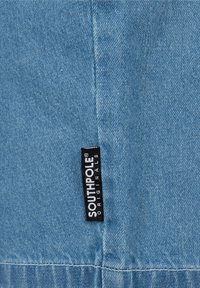 Étiquette en tissu noir avec "SOUTHOLE ORIGINALS" en blanc, attachée à un tissu en denim bleu clair avec des coutures visibles.