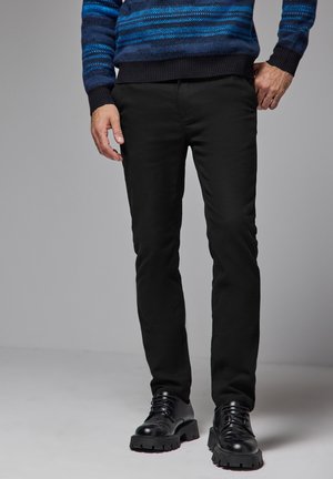 Pantalon slim noir avec une texture lisse, associé à des chaussures noires brillantes. Le modèle porte un pull rayé bleu.