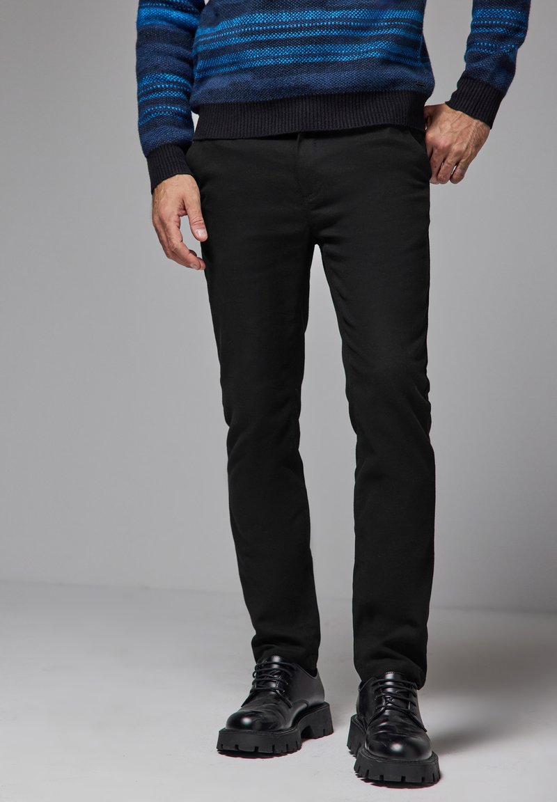 Svarta slim-fit byxor med en slät yta, kombinerat med glänsande svarta skor. Modellen bär en blårandig tröja.