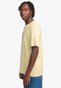 Jonge man met korte twists, gekleed in een lichtgele graphic t-shirt en blauwe jeans, staande zijwaarts tegen een witte achtergrond.