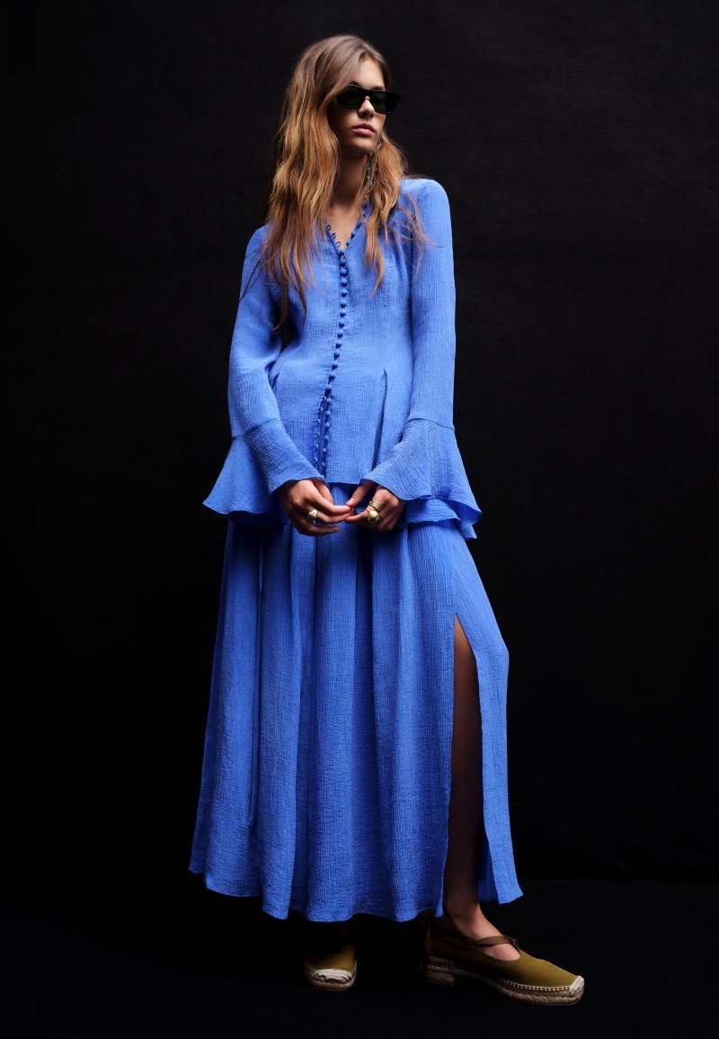 Γυναίκα με μακρύ μπλε υφασμάτινο φόρεμα με κουμπιά μπροστά, φαρδιά μανίκια και ψηλή σχισμή, φορώντας σκούρα γυαλιά ηλίου και πράσινα εσπαντρίγιες.