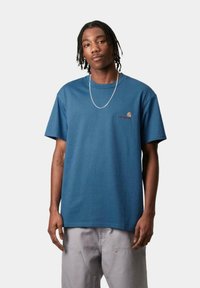 Camiseta de algodón azul con cuello redondo, mangas cortas y un pequeño logo bordado en el pecho, combinada con pantalones gris claro.