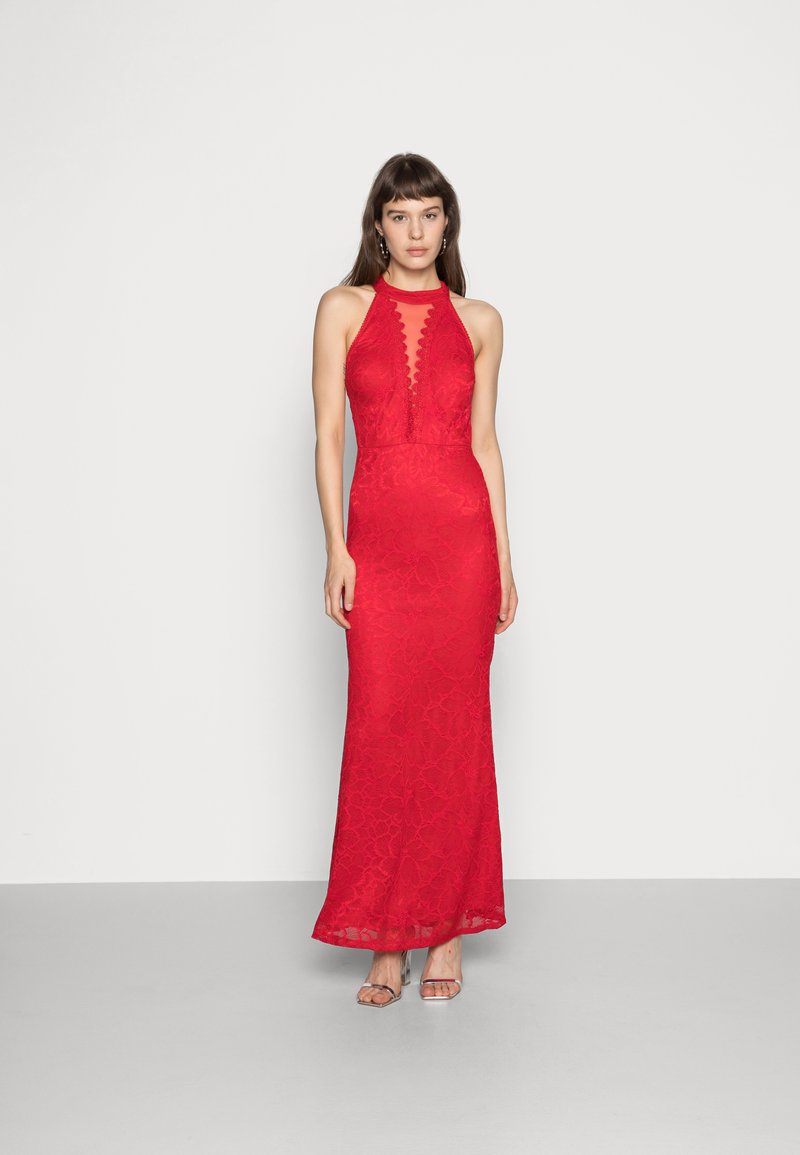 Vestido de noite vermelho em renda com silhueta ajustada, decote complexo em forma de escalope e barra até ao chão. Modelo a usar sandálias de salto transparente.