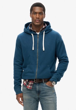 Blue zip-up hoodie with light drawstrings, soft fabric, front pockets, worn over a plaid shirt, paired with jeans and a watch.