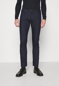 Mörka denimjeans med slim fit, femfickorsdesign, kontrasterande sömmar och klassisk knappstängning, kombinerat med svarta skor.