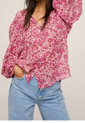 Frau trägt eine rosa geblümte Bluse mit langen Puffärmeln und hellblauen Jeans mit hoher Taille, eine Hand in der Tasche.