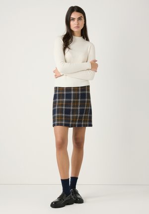 Weißes, geripptes Langarmoberteil, kombiniert mit einem marineblauen Karo-Rock mit orangefarbenen und weißen Akzenten. Das Modell trägt schwarze Slipper und blaue Socken.