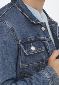 Sininen farkkutakki, jossa on klassinen kaulus, kaksi etutaskua nappisuljennalla ja kontrastiväriset ompeleet. Rakenteellinen kangas.