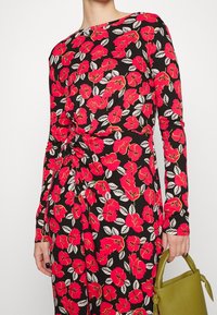 Kvinna klädd i en långärmad svart klänning med lysande röd blommönster och håller en liten olivgrön handväska i sin högra hand.