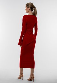 Abito midi rosso a costine con maniche lunghe, polsini a campana e uno spacco sul retro; abbinato a sandali con tacco alto e fasce nude. La texture è liscia.