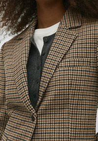 Blazer à carreaux marron avec un col cranté, présentant des motifs complexes. En dessous, un cardigan boutonné gris foncé est visible.