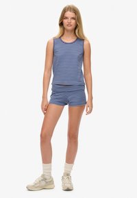 Jonge vrouw die staat en een blauwe en grijs gestreepte mouwloze top en bijpassende shorts draagt, met witte sokken en beige sneakers.