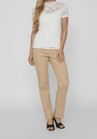 Vit spetsblus med korta ärmar och blommönster, kombinerad med beige raka byxor. Modellen bär svarta avslöjande klackar.