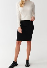 Cremfarbener Pullover mit langen Ärmeln, kombiniert mit einem enganliegenden schwarzen Rock in Knielänge und schwarzen Schnürschuhen. Weiches Material mit glatter Textur.