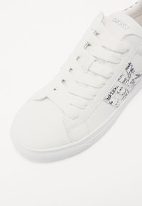 Sneaker bianca DKNY realizzata in materiale sintetico liscio. Presenta lacci piatti, una suola in gomma testurizzata e un pannello laterale con un motivo in bianco e nero.