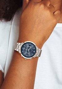 Montre-bracelet en métal argenté avec un cadran rond bleu marine, trois sous-cadrans et un bracelet à maillons poli. Accents en forme de T sur le boîtier.