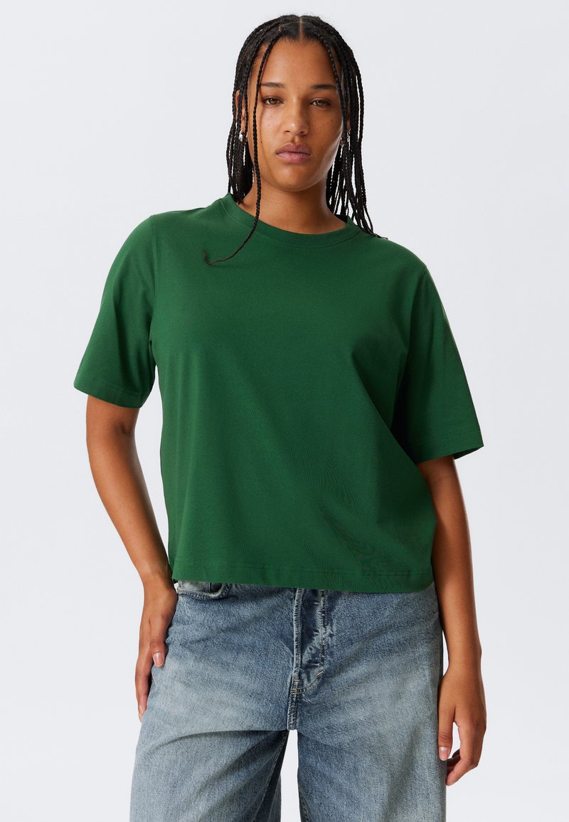 T-shirt in cotone verde con una vestibilità rilassata, collo tondo e maniche corte, abbinata a jeans azzurri a taglio largo.
