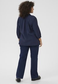 Navyblaue strukturierte Bluse mit gepufften Ärmeln, leicht gerafft am Hals. Kombiniert mit dunkelblauen Schlagjeans und schwarzen Schuhen.