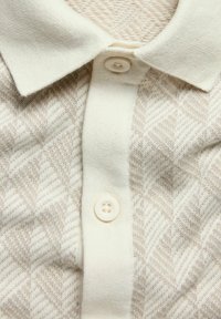 Crème-kleurige polo met een gestructureerd visgraatpatroon, voorzien van een klassieke kraag en een knoopsluiting bij de hals.
