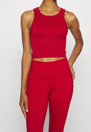 Kobieta ubrana w czerwony krótki top bez rękawów i czerwone legginsy z wysokim stanem na jednolitym tle.