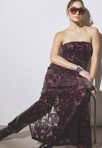 Brezkrajna, dolga obleka v temno vijoličnih bleščicah z cvetličnim čipkastim premazom. Manekenka nosi velike sončna očala in zapestnico s členki; črne visoke škornje do stegen.