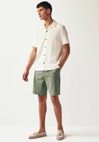 Chemise crème à manches courtes avec texture et boutons marron, portée avec un short vert et des chaussures à enfiler beiges avec des accents décoratifs.