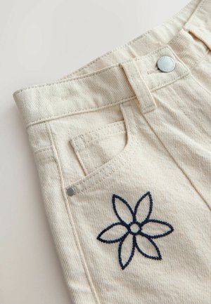 Gros plan sur la poche d'un pantalon en denim beige avec un bouton argenté et un détail brodé d'une fleur à six pétales bleu marine.