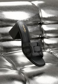 Schwarze High-Heel-Sandale zum Hineinschlüpfen mit breitem Riemen und quadratischer Zehenpartie, die auf einer glänzenden metallisch-silbernen gepolsterten Oberfläche ruht.