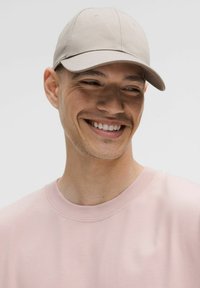 Hombre con una gorra beige y una camisa rosa clara, sonriendo y mirando hacia un lado sobre un fondo claro y liso.