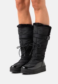 Pitkät mustat talvikengät, joissa on turkisreunus, tikattu kangas, nauhakiinnitys edessä, sivuvetoketju ja paksu kumipohja, käytettynä paljaille jaloille.