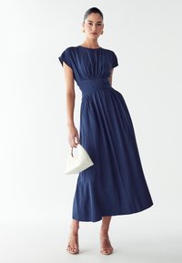 Temno modra maxi obleka z nabranim pasom, kratkimi rokavi in gladko teksturo. Združena z belo torbico in nude zapetimi čevlji.