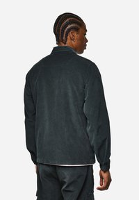 Giacca in corduroy verde scuro con taglio dritto, maniche lunghe e colletto a banda. Tessuto sottile e polsini elasticizzati.