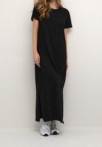 Robe longue noire à manches courtes en tissu lisse avec une fente latérale, associée à des baskets blanches et grises. Coupe simple et décontractée.