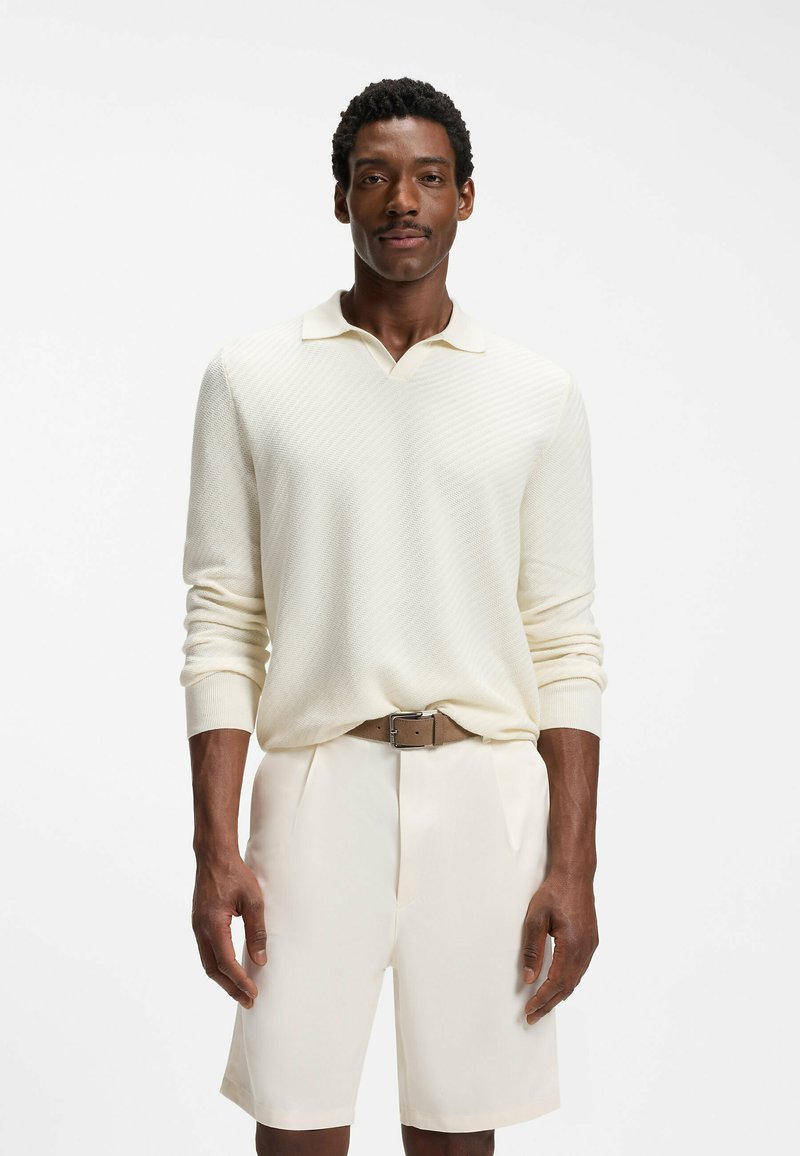 Homme portant un pull polo à manches longues texturé crème, un short crème et une ceinture marron, debout devant un fond blanc uni.