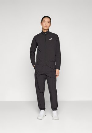 Zwarte sportieve zip-up jas met een staande kraag, bijpassende broek en witte sneakers. De jas heeft een Puma-logo op de borst.