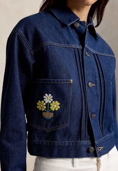 Jachetă din denim cu nasturi în față și un buzunar mare decorat cu flori galbene și albe din mărgele, într-un ghiveci maro.