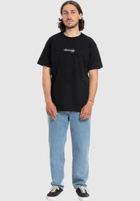 Zwarte katoenen t-shirt met grafische opdruk "HomeFly" in wit, ruime pasvorm, ronde hals. Gecombineerd met lichtblauwe jeans en zwarte sneakers.