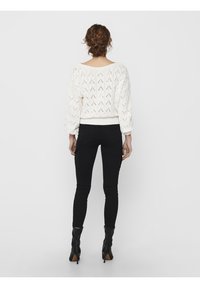 Weißer Strickpullover mit lockerer Passform und gemusterter Textur; kombiniert mit schwarzen Slim-Fit-Hosen und schwarzen Stiefeletten.