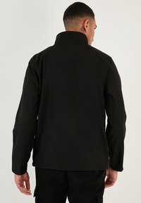 Chaqueta negra con un cuello alto, hecha de tela suave. Presenta mangas largas y un corte recto. Destaca por su diseño minimalista y la ausencia de herrajes visibles.