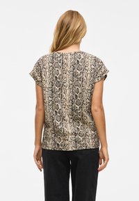 Frau mit langen blonden Haaren, die ein kurzärmliges Oberteil mit Schlangenmotiv und schwarze Hose trägt, steht mit dem Rücken zu einer neutralen Hintergrund.
