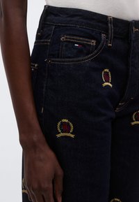 Tumedast denimist teksapüksid kollaste õmblustega, millel on tikitud vimplimustrid punases, rohelises ja kuldses värvitoonis. Klassikaline viie taskuga disain.