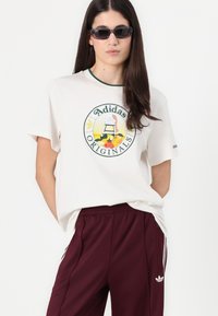 Tricou din bumbac bej deschis cu un logo grafic rotund care înfățișează un coș cu fructe; asociat cu pantaloni atletici vișinii și detalii albe Adidas.