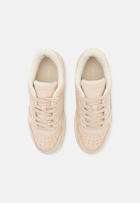 Ett par beiga Lacoste-sneakers sedd ovanifrån med snören, perforerade tåskydd och vadderade kragar på en vit bakgrund.