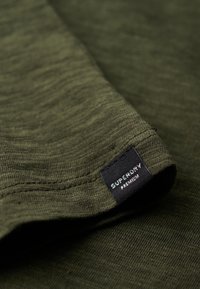 Tejido de algodón de color verde oliva con una textura suave, que presenta un borde doblado y una etiqueta negra con "SUPERDRY PREMIUM" impresa en blanco.