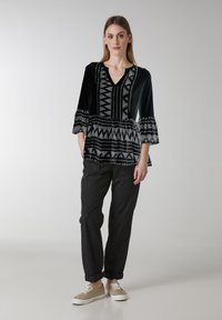 Top peplum bianco e nero con scollo a V, maniche a campana e motivi tribali a contrasto. Indossato con pantaloni neri e sneakers beige.